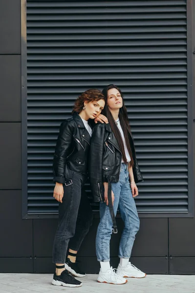 Portrait of two beautiful models on a dark wall background, wearing stylish casual clothes, posing for the camera.Two buttresses are standing outside on a dark background and looking at the camera — 스톡 사진