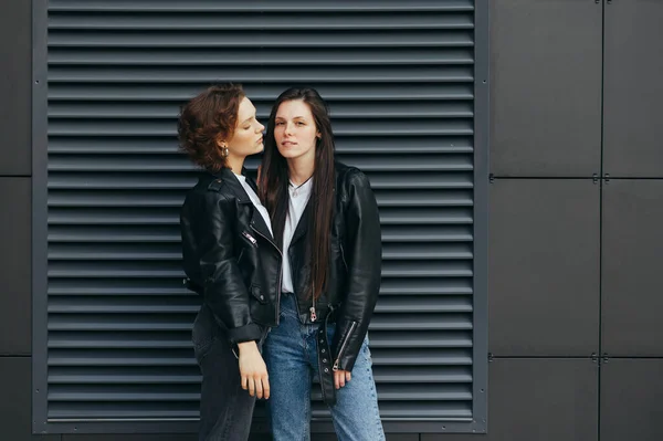 Retrato de duas meninas sexy em jaquetas de couro, de pé sobre um fundo de parede escura e posando para a câmera. Par de lésbicas fica em fundo preto. Conceito de orgulho. Bela namorada no fundo . — Fotografia de Stock