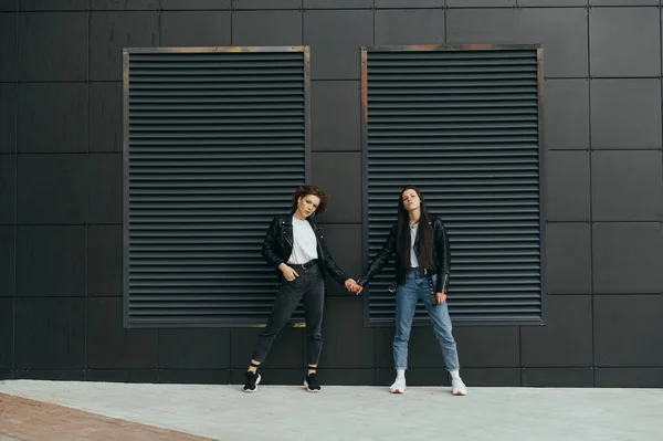 Two stylish girls are holding hands against a black wall, posing for the camera. Portrait of two attractive models in leather jackets standing on black background holding hands. Copy space — 스톡 사진