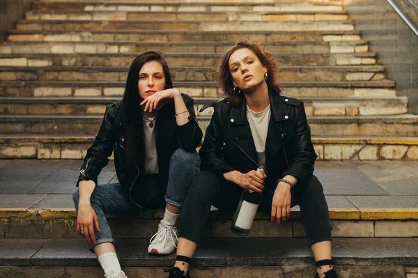 Portrait of two girls models in stylish casual clothes sitting on stairs with bottle of wine in hand posing at camera with serious face. Two girlfriends are sitting on the stairs in the underground — 스톡 사진