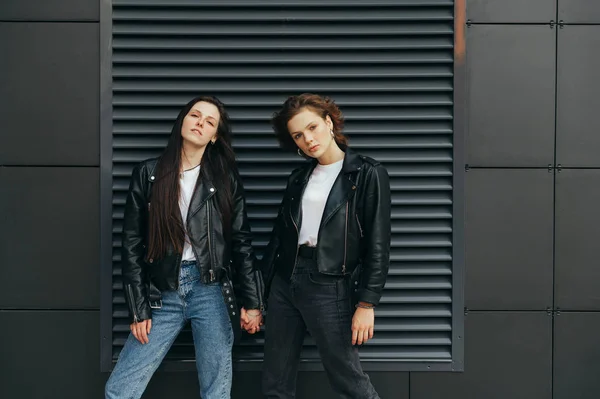 Street portrait of two stylish girls in leather jackets are standing against a dark wall, holding hands, posing for the camera with serious faces. Photo of lesbian models on dark background — 스톡 사진