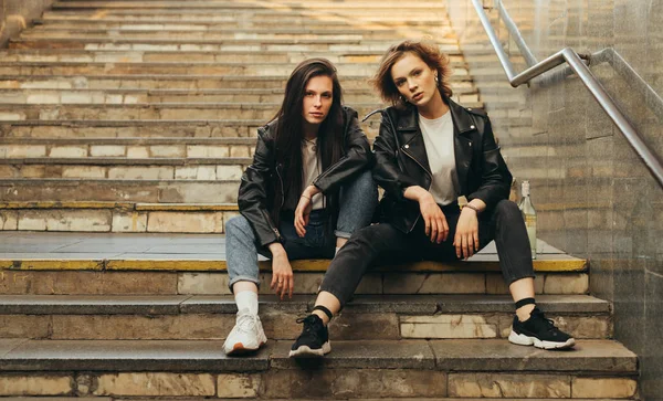 Photo of two models on the stairs of the subway,focused looking at the camera. Two girlfriends in casual clothes,wearing leather chicken, sitting on the stairs and posing on camera with a serious face — 스톡 사진