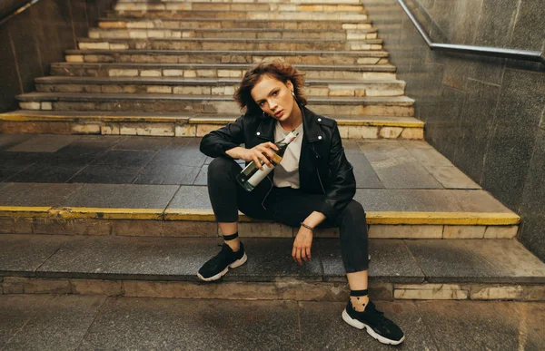 Retrato de uma menina apta em uma jaqueta de couro senta-se nas escadas do metrô com uma garrafa de água na mão, olha na câmera com uma cara séria.Foto modelo elegante na pose subterrânea — Fotografia de Stock