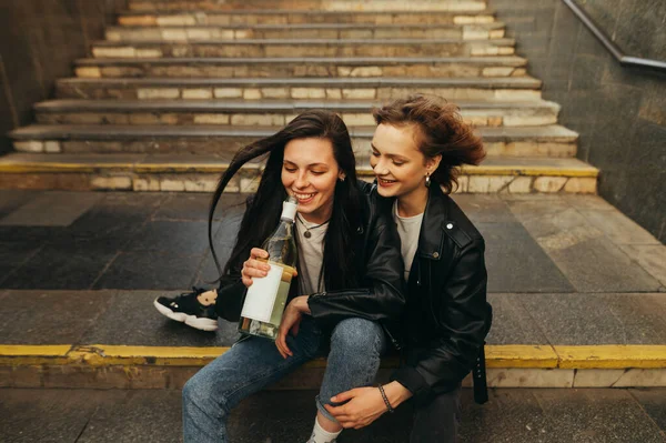 Дві щасливі дівчата в стильному одязі сидять на сходах під землею, п'ють вино і розважаються з посмішками. Подружки свободи п'ють вино з пляшки на вулиці, сидячи на сходах — стокове фото