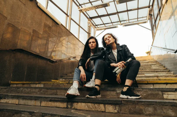 Portrait of two models in stylish casual clothes sitting on stairs with bottle of wine in hand posing at camera with serious face. Two girls are sitting on the stairs in the underground — 스톡 사진