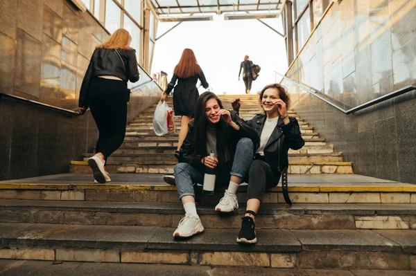 Zwei fröhliche Mädchen, die mit einer Flasche Wein auf der Treppe in der U-Bahn sitzen und lächeln. stilvolle Freundinnen reden und lachen, trinken Wein in der U-Bahn vor dem Hintergrund einer Passantin. — Stockfoto
