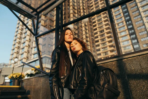 Street fashion portrait of two girls in leather jackets on urban background, looking at camera and posing with serious face.Attractive girlfriends standing on the stairs on an architecture background — Stock Photo, Image