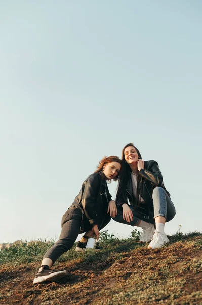 Duas meninas positivas em roupas casuais elegantes posando na colina contra o fundo do céu, sorrindo e olhando para a câmera. Meninas felizes em jaquetas de couro agachadas na colina e se divertindo . — Fotografia de Stock