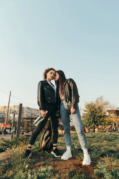 两个穿着街头服装的女孩站在草地上，站在城市景观的背景上，在镜头前摆姿势的时尚画像。街上穿着皮夹克的迷人模特的画像。垂直方向 — 图库照片