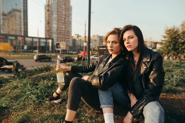 Portrait deux copines branchées assises le soir sur le sol avec une bouteille de vin à la main sur fond de paysage urbain et posant à la caméra.Deux filles modèles se sont assis pour se reposer sur l'herbe pour une promenade — Photo