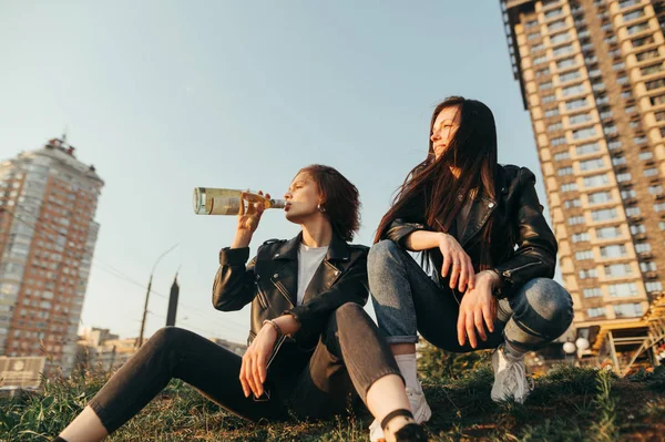Portré két elegáns barátnő ül egy dombon az égen háttér iszik bort naplementekor. Két lány sétál bort iszogatva és pihentetve a földön ülve a városkép hátterében.. — Stock Fotó