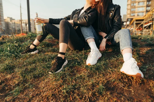 Háttér.Közelkép fiatal leszbikus lányok lábait ül a földön egy üveg bor a kezükben, és ölelés.Lábak a lányok tornacipőben ül a füvön a városkép háttér naplementekor — Stock Fotó