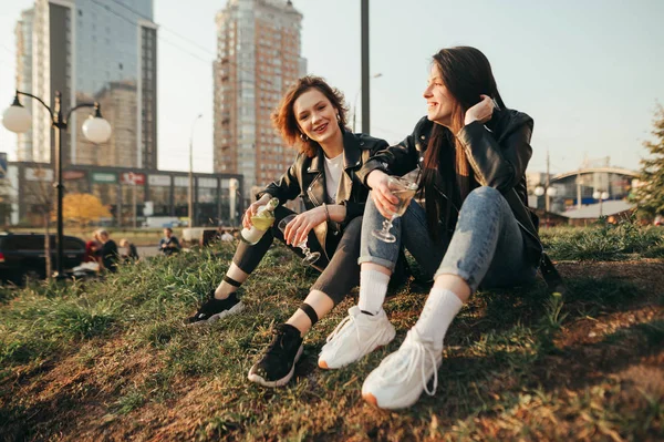 Porträt glücklicher junger Mädchen in Lederjacken, die auf dem Gras vor dem Hintergrund der Stadtlandschaft sitzen, mit einer Flasche Wein und Gläsern in den Händen, die in die Kamera lächeln. Freundinnen trinken Wein beim Spaziergang. — Stockfoto