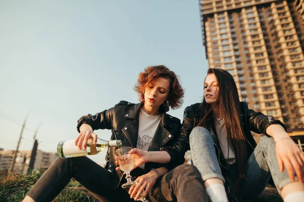 A lány bort önt palackból üvegbe az utcán, városi háttérrel. Stílusos barátnők ülnek a füvön az égen és az építészet háttérben és bort isznak. A lányok romantikusan isznak alkoholt.. — Stock Fotó
