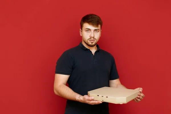 Портрет человека с бородой на красном фоне с коробкой пиццы в руках, смотрит в камеру. Бородатый молодой человек с коробкой пиццы с коробкой доставки в руках изолированы на красном фоне — стоковое фото