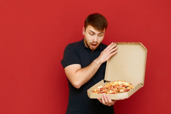 Koyu renk tişörtlü şaşkın adamın portresi kırmızı arka planda duruyor, açık kutuya pizza ile şaşırmış gibi bakıyor. Şok olmuş suratlı aç sakallı adam taze pizzaya bakıyor. Boşluğu kopyala — Stok fotoğraf