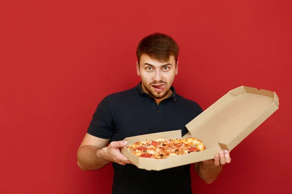 Sakallı komik bir adam teslimatta pizza kutusunu tutuyor ve aç ve şehvetli bir yüzle kameraya bakıyor. Kırmızı arka planda pizza kutusu olan aç bir adamın portresi. Fast food konsepti. — Stok fotoğraf
