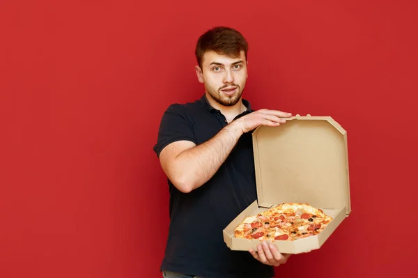 Pozitif bir adamın portresi kırmızı arka planda duruyor ve bir kutu lezzetli taze pizzayı açıp kameraya bakıyor. Adam elinde bir kutu taze pizza tutuyor ve kamerayı işaret ediyor. İzole edilmiş. Boşluğu kopyala — Stok fotoğraf