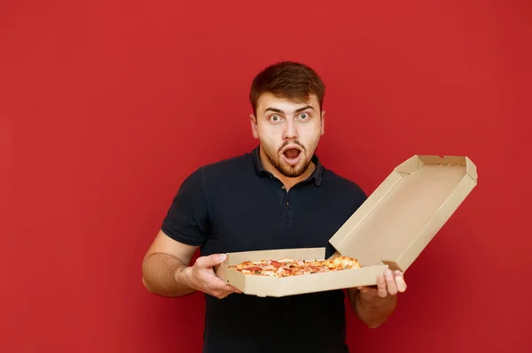 Sakallı genç adam elinde sıcak pizza kutusuyla kırmızı arka planda şok olmuş bir şekilde kameraya bakıyor. Elinde lezzetli pizzayla şaşırmış aç bir adamın portresi. Fast food. — Stok fotoğraf