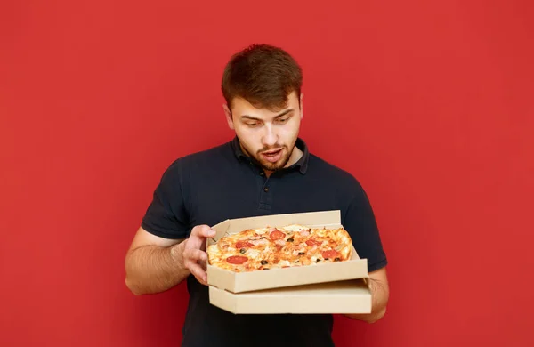 Bearded man in a black T-shirt is holding a box of fresh pizza and staring at a fast food on a red background with a hungry look. Portrait of hungry man going to eat hot pizza out of the box. — Stock Photo, Image