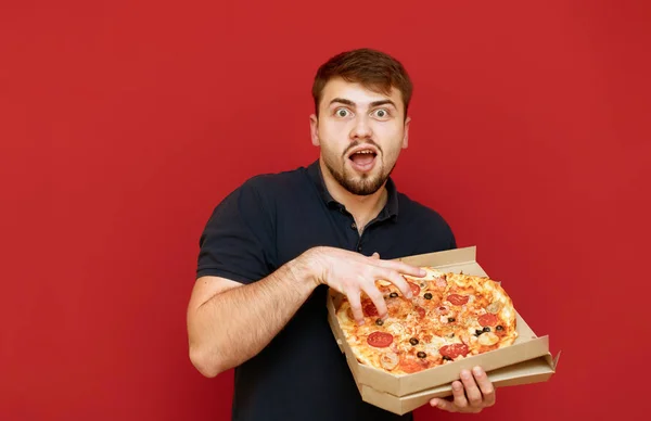 Портрет експресивної людини в темній футболці на червоному тлі, тримає коробку зі свіжою піцою в руці, дивиться в камеру з божевільним обличчям і бере шматок. Ізольовані — стокове фото