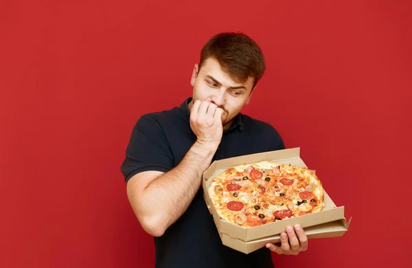 Человек стоит на красном фоне с коробкой горячей пиццы и смотрит на еду и кусает пальцы от нервов. Человек с диетой держит в руке открытую коробку вкусной пиццы. Вредная пища — стоковое фото