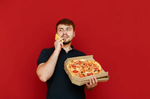 Портрет улыбающегося молодого человека в черной футболке и с бородой, выделенной на красном фоне, с коробкой пиццы в руках, разговаривающего по телефону, отворачивающегося и улыбающегося. Мужчина приглашает друзей на горячую пиццу — стоковое фото