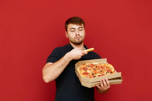 Чоловік стоїть на червоному тлі з піцою коробкою в руці і робить фото піци на смартфоні для інтернету. Молодий чоловік фотографує піцу при доставці. Блогер робить фотографію піци. Відокремлені. — стокове фото