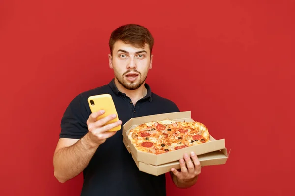 Sakallı bir adam kırmızı bir arka planda elinde pizza kutusu ve elinde akıllı bir telefonla şaşırmış bir yüzle kameraya bakıyor. Adam internetten pizza sipariş etmiş.. — Stok fotoğraf