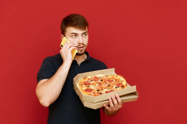 Sakallı adam elinde bir kutu pizzayla kırmızı arka planda duruyor ve telefondaki yüzükler ciddi bir yüz ifadesiyle yüzünü çeviriyor. Adam telefonla pizza sipariş ediyor.. — Stok fotoğraf
