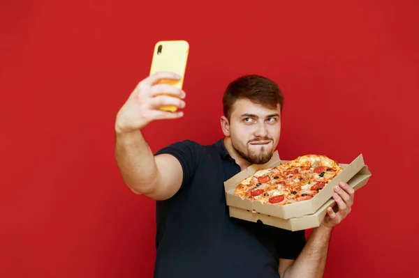 赤い背景にピザの箱で自撮りした面白い髭の男は、スマートフォンを見て面白い顔をしています。ピザの宅配ボックスで写真を撮る陽気な男。隔離。. — ストック写真