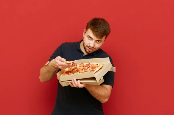 Komik sakallı adam, kırmızı bir arka planda pizza dilimini kutudan çıkarıp kameraya vahşice bakıyor. Siyah tişörtlü aç adam, kutudan lezzetli bir sıcak pizza yiyor. — Stok fotoğraf