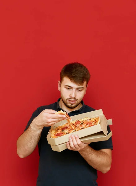 Homem engraçado com fome fica em um fundo vermelho e tira uma fatia de pizza da caixa. Um jovem engraçado com barba a comer pizza fresca da caixa. Isolado. Fast food . — Fotografia de Stock