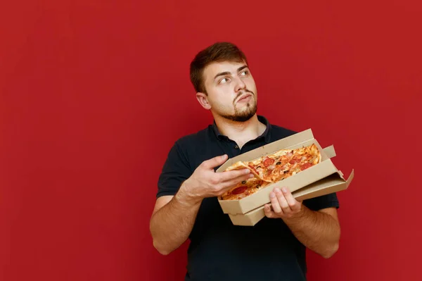 El hombre con una caja de pizza se para sobre un fondo rojo, toma una rebanada de comida rápida y mira hacia otro lado con una cara seria. Hombre barbudo hambriento comiendo pizza caliente. Aislado. Copiar espacio — Foto de Stock