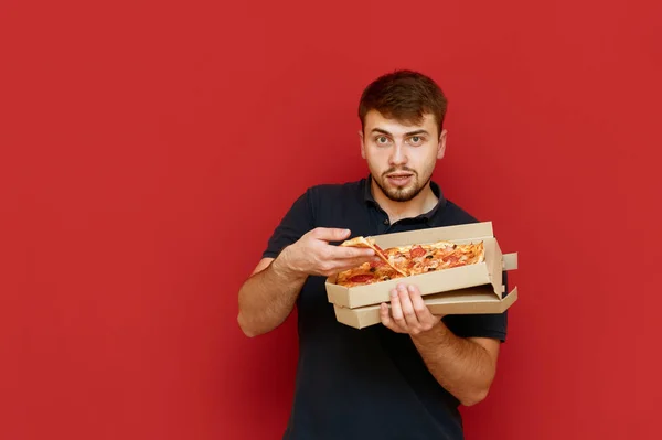 Elinde bir kutu pizzayla kırmızı arka planda duran sakallı bir adamın portresi kameraya bakar ve kutudan bir dilim pizza alır. Fast food yiyen aç bir adam. İzole edilmiş. — Stok fotoğraf