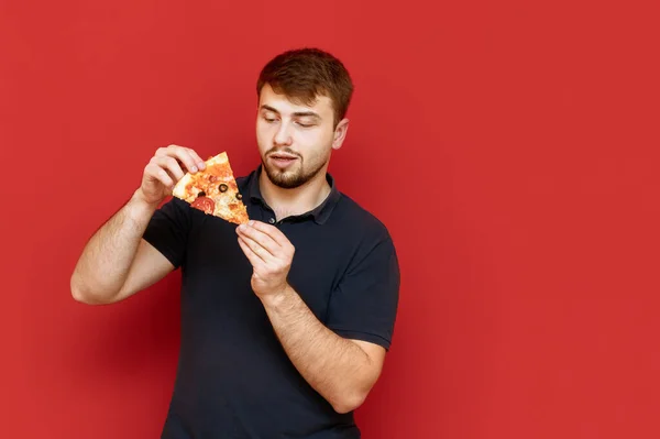 Homem faminto de t-shirt preta e com barba isolada em fundo vermelho com um pedaço de pizza nas mãos, com fome. Retrato de um homem com uma fatia de pizza, isolado. Fast food . — Fotografia de Stock