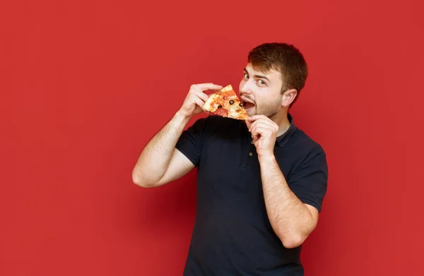 Porträt eines fröhlichen hungrigen Mannes mit Bart, der Pizzascheiben auf rotem Hintergrund isst, in die Kamera schaut und lächelt. Bärtiger Mann beißt isoliert in ein Stück Pizza. — Stockfoto
