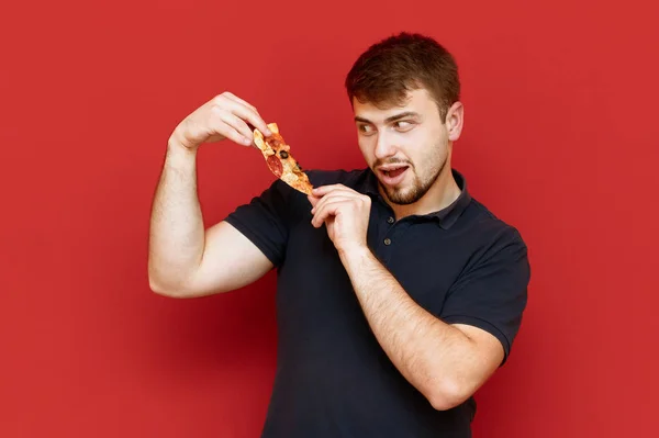 Голодный бородатый мужчина играет с кусочком пиццы в руках на красном фоне и улыбается. Веселый человек держит кусок пиццы и смотрит на фаст-фуд с голодным взглядом. Isolated . — стоковое фото