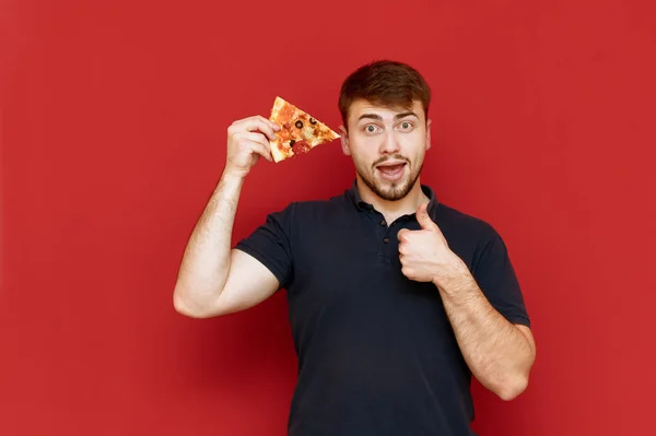 Позитивный человек с бородой стоит на красном фоне с кусочком пиццы в руке, смотрит в камеру, улыбается и показывает большой палец вверх. Голодный парень позирует с кусочком пиццы и показывает жест симпатии — стоковое фото