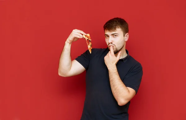 Портрет веселого бородатого мужчины с вкусным ломтиком пиццы в руках на красном фоне, смотрит в камеру, улыбается и облизывает палец. Человек наслаждается вкусной пиццей, съедает последний кусочек . — стоковое фото