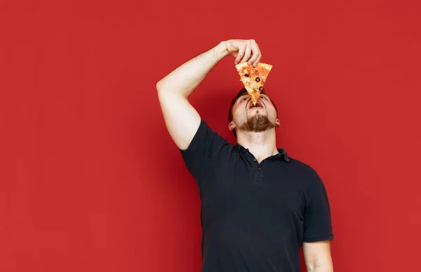 Un hombre gracioso con una camiseta oscura comiendo pizza sobre fondo rojo, levantando la cabeza y metiéndose un trozo en la boca. Foto del hombre comiendo la última rebanada de pizza, aislado. Comida rápida. Copiar espacio — Foto de Stock