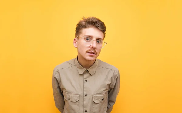 Retrato de um lindo nerd com bigode e óculos fica contra um fundo amarelo e olha atentamente para a câmera, vestindo uma camisa bege. Engraçado estudante masculino na camisa isolada no amarelo — Fotografia de Stock