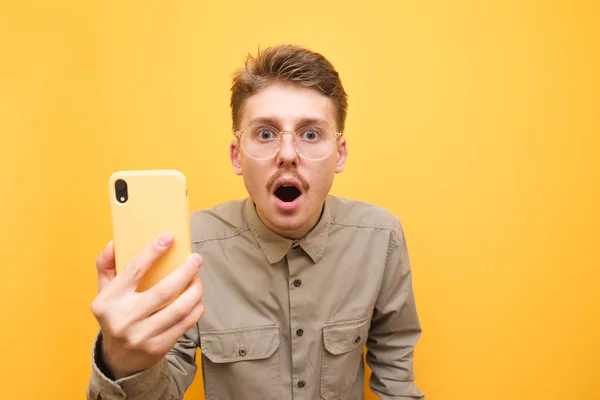 Retrato de nerd sorprendido en gafas y con bigote sobre fondo rojo, mira a la cámara con la cara impactada y sostiene un teléfono inteligente en las manos, usando camisa beige. Aislado . — Foto de Stock