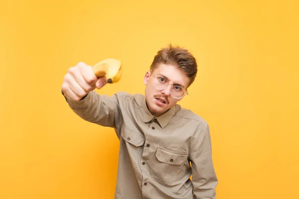 Смешной парень в очках и рубашке направляет банановый пистолет на камеру и смотрит с серьезным лицом на желтом фоне. Портрет ботаника с усами, держащего в руке банановое оружие. — стоковое фото