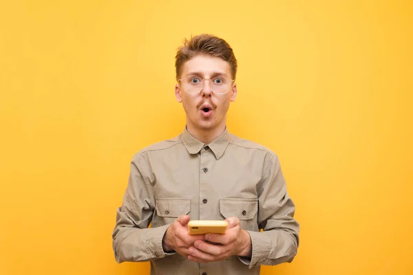 Joven sorprendido en gafas y camisa y bigote se coloca en el fondo amarillo con el teléfono inteligente en la mano, mira a la cámara con la cara sorprendida. Retrato de estudiante nerd sorprendido con smartphone . — Foto de Stock