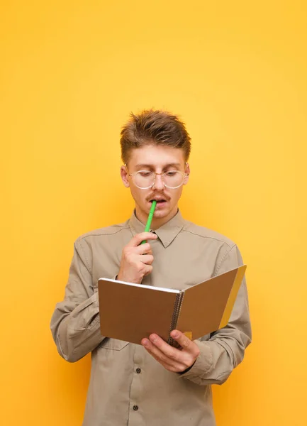 Retrato de un hombre nerd joven reflexivo de pie sobre un fondo amarillo con un cuaderno en la mano, mordiendo un bolígrafo y mirando pensativamente en un libro de trabajo. Estudiante reflexivo hace la tarea . —  Fotos de Stock