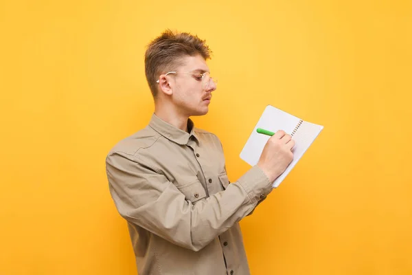 Retrato de un estudiante con camisa y gafas sobre un fondo amarillo y escribe un bolígrafo en un cuaderno. Tipo serio nerd realiza tareas estudiantiles. Concepto de estudiante. Copiar espacio — Foto de Stock