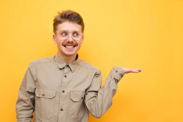 Glad nörd i glasögon och skjorta står på en gul bakgrund, tittar bort och pekar handen på kopieringsutrymme. Rolig glad kille med mustasch presenterar ett tomt utrymme för text med handen och leenden — Stockfoto