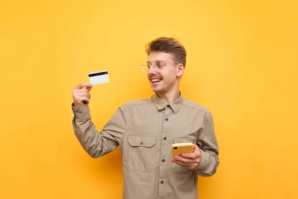 Ritratto di giovane felice con smartphone e carta di credito in mano su sfondo giallo, guardando carta di credito e sorridente. Il ragazzo felice sta facendo shopping online. Copia spazio — Foto Stock