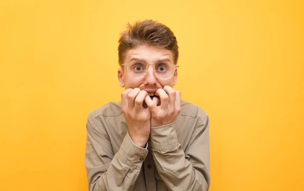 Retrato de un joven asustado con camisa y gafas sobre fondo amarillo, mira a la cámara con la cara conmocionada y se muerde los dedos. Nerd asustado aislado sobre fondo amarillo. Copiar espacio — Foto de Stock